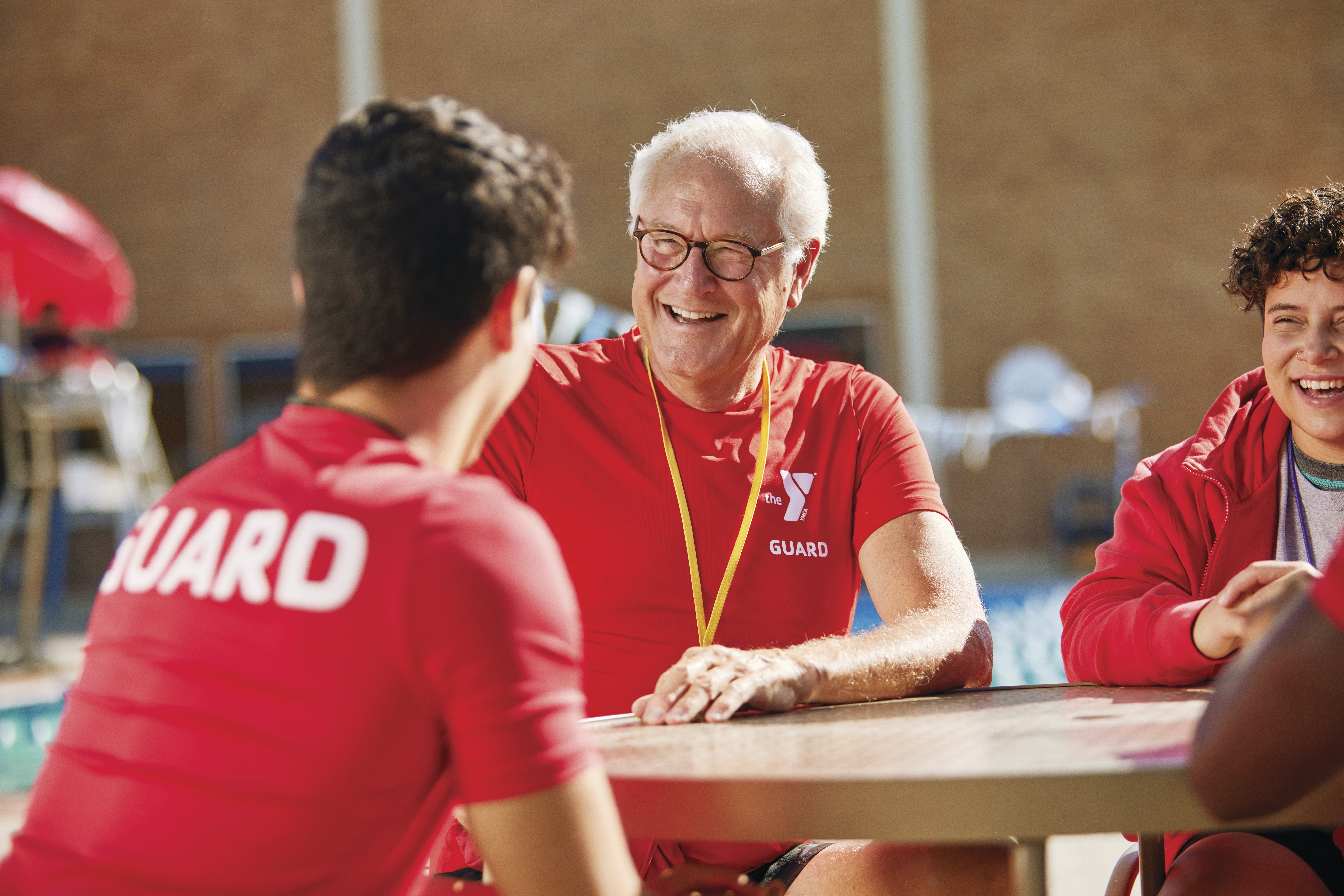 YMCA lifeguards sit outside by the pool around a table and during training talking and smiling.